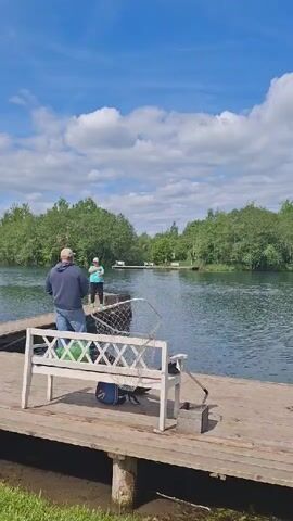 The video shows two people fishing on a wooden pier next to a body of water, likely a lake or large pond, in Kipen, Russia.

**Subjects:**
In the foreground, a person wearing a blue hoodie and jeans stands on the wooden pier, holding a fishing rod. Behind them, a white wooden bench, a fishing net, a blue bag, and a metal bucket are visible on the pier. Further down the pier, another person, wearing a light-colored shirt and possibly a hat, is also fishing.

**Scene and Setting:**
The wide body of water stretches into the background, with lush green trees and foliage lining its banks. Another small wooden structure, possibly a pier or small dock, can be seen in the distance across the water. The sky is bright blue with scattered white, fluffy clouds, indicating good weather.

**Activity:**
Both individuals appear to be engaged in fishing. The person in the blue hoodie seems to be actively casting or retrieving their line, while the other person is also holding a fishing rod, suggesting they are waiting for a bite or reeling in.

**Notable Details:**
The scene depicts a peaceful and recreational outdoor activity on a bright, sunny day. The presence of fishing equipment and the natural environment reinforce the theme of leisure and angling. There is no visible text in the video.