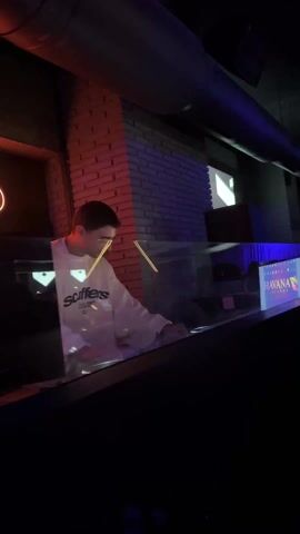 The video shows the inside of a nightclub. The opening shots are shaky and blurred, focusing on a person raising their arms in excitement amidst a crowd of people. Blue and red lights pulse in the background, illuminating the space with an energetic, party atmosphere. The video then focuses on a DJ, standing behind a clear barrier, operating electronic equipment on a DJ console. The DJ is a young man, wearing a white sweater, focused on mixing music. A neon sign illuminates the wall behind him, reading "lló".