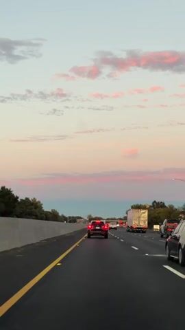 The video shows a view from a car driving on a highway at dusk. The sky is filled with soft pink and blue clouds, indicating sunset. Several cars and a large truck are visible on the road ahead, all traveling in the same direction. The highway has a concrete barrier on the left and a double yellow line marking the lanes. Trees and vegetation line the side of the highway. The overall scene depicts a typical highway commute during a colorful sunset.