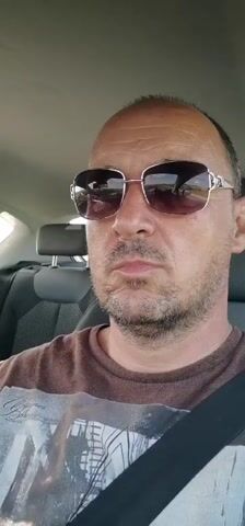A man wearing sunglasses and a brown t-shirt sits in the driver's seat of a car, looking at the camera. The background shows the car's interior, including the headrest and seatbelt. The video appears to be a selfie taken while driving.