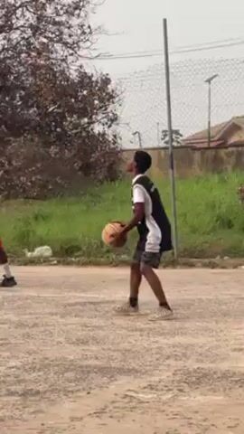 The video shows four young men playing basketball on an outdoor court. Three of the young men are wearing red jerseys and shorts, and the fourth is wearing a white-and-black jersey and dark shorts. The player in the white-and-black jersey dribbles the basketball, takes a step back, and shoots toward the basketball hoop. The court surface is dirt. The background shows overgrown grass, shrubs, trees, and a chain-link fence. A utility pole is visible to the right of the fence.
