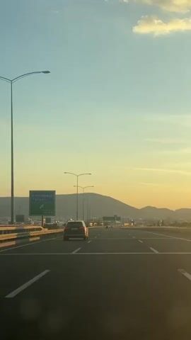 A static shot from within a car traveling on a highway at dusk. The sky is a gradient of pale blue to light orange near the horizon. The road ahead is straight with multiple lanes, and several cars are visible in the distance, the closest being a dark-colored SUV directly ahead. Streetlights line the highway. A green highway sign reads "Kemalpaşa Torbalı 400m". In the background, a silhouette of a hill or mountain can be seen.