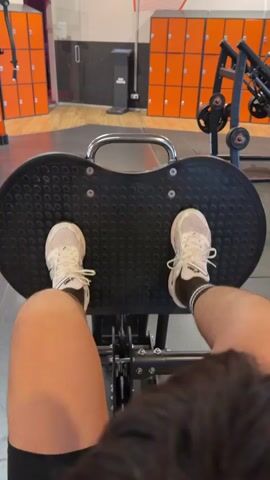 The video shows a first-person view of a person performing a leg press exercise on a machine. The person's legs, clad in white sneakers and black socks, are positioned on a large black platform with a textured surface. The knees are bent in the initial phase of the exercise and then extend outward. In the background, orange lockers are visible, suggesting the setting is a gym. The lighting is bright, and the overall atmosphere appears to be one of physical activity.