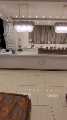 The video shows the interior of a store called "Rest & Sweets." The camera focuses on a long white counter with gold trim. Behind the counter, there are several decorative items including various metallic teapots. A person wearing black clothes walks in front of the counter, partially obscuring the view. Overhead, there are two large chandeliers with gold and crystal accents. The floor is tiled with large square tiles. The store appears to be well-lit and decorated in a modern style.