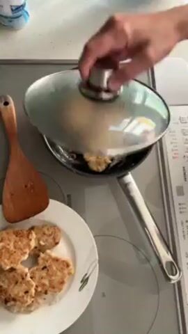 A fair-skinned woman wearing a black tank top and a beaded necklace cooks in her kitchen. The camera points down at a stovetop with a stainless steel frying pan where four small, round fritters sizzle in oil. A wooden spatula sits beside the pan. A white plate with green trim holds more fritters already cooked. The stovetop is white with digital controls on the right. The woman appears in the frame and smiles slightly at the camera. She moves around the kitchen with the camera following her, but her actions are not clearly visible.