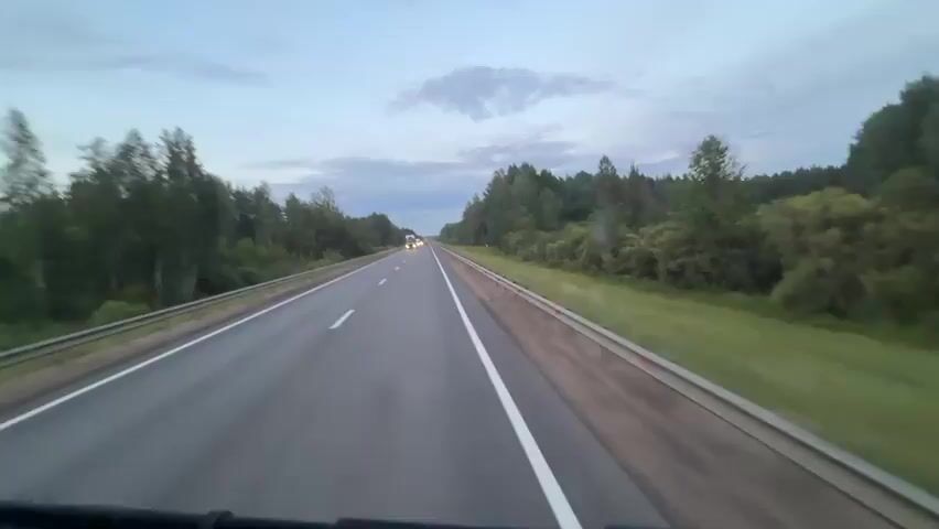 The video shows a point-of-view shot from inside a vehicle driving on a highway. The sky is overcast and the lighting suggests it is either dawn or dusk. The highway has two lanes in each direction separated by a solid white line. A metal guardrail runs along the left side of the road. On both sides of the highway, there are dense forests with tall trees. In the distance, other vehicles, including trucks, can be seen on the highway. One white truck with headlights on passes the camera's vehicle. The camera is looking forward, and the bottom edge of the vehicle's dashboard is visible.
