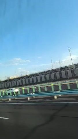 The video shows a view from a moving vehicle on a highway. The highway has multiple lanes with a green and white barrier. On the right side of the highway, there is a concrete retaining wall, possibly part of an elevated road or structure, with power lines and poles visible above it. The sky is blue with some clouds. Several cars are seen driving in the same direction as the camera. The lighting suggests it is daytime, possibly late afternoon or early morning due to the angle of the light. The motion blur indicates the video was captured while in motion.