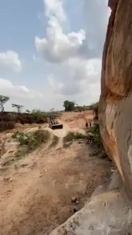 The video shows a yellow front-end loader maneuvering in what appears to be a mining or excavation site. The ground is sandy and uneven, with some vegetation and a large rock formation visible on the right side of the frame. Several people can be seen in the background, some standing and others sitting. The sky is partly cloudy. The loader moves forward, navigating the rough terrain.