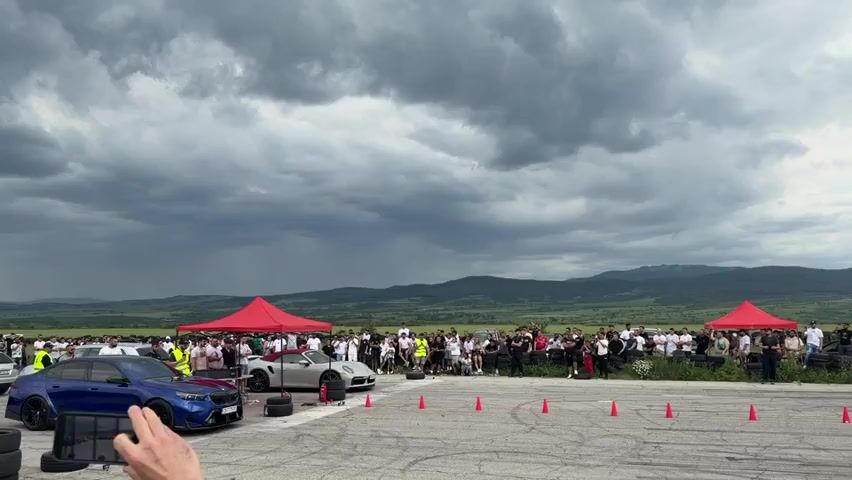 The video captures a drag race event. A crowd of spectators lines either side of the race track, which is marked by orange cones. Two cars, a blue sedan and a silver convertible, are at the starting line. The race begins, and the cars accelerate down the track. In the background, a landscape of rolling hills and a cloudy sky can be seen. Red tents are set up on both sides of the track.