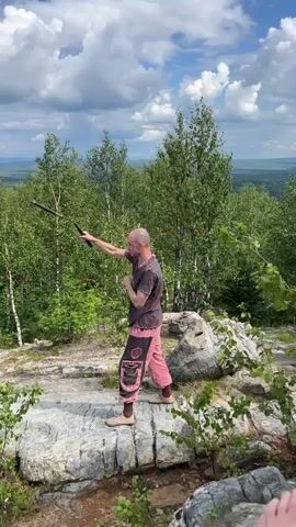 A bald man wearing a dark t-shirt and loose pink pants with black patterns on them is practicing a martial art or dance on a rocky outcropping. He is holding a dark object that appears to be a bo staff or a similar prop. The man is positioned with his back to the camera and is in the midst of a movement, with one arm raised and the other extended downwards. He is surrounded by lush green trees, and in the background, a hazy landscape stretches out under a partly cloudy sky. The rocks he stands on are large and grey, with some smaller green plants growing around them.