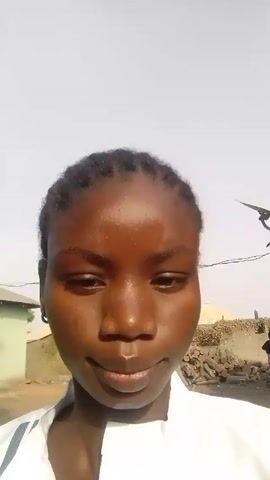 A young woman with braided hair looks directly at the camera with a slight smile. She wears a white top. The background shows a rural setting, with a small green building, a pile of logs, and a tree visible under a bright sky. The video appears to be taken outdoors in daylight.