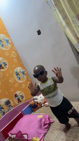 A short video shows a toddler in sunglasses standing in a room decorated for a baby shower. The toddler is wearing a white tank top with black shorts and is barefoot. He's standing on a bed with toys nearby. The wall behind him is decorated with a Smurfs wallpaper. There's also a "Welcome Baby" banner hanging above a curtain. The toddler is waving his hands and making sounds.