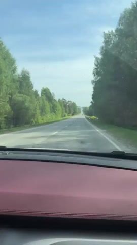 A car travels on an empty, two-lane road under a partly cloudy, blue sky. The road is straight and long, stretching out to the horizon. Lush green trees line both sides of the road. A metal guardrail is visible on the right side of the road. The camera is positioned inside the car, behind a dashboard with a red surface. The car appears to be in motion, as indicated by the blurring of the scenery.