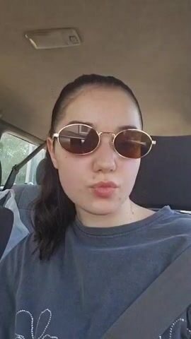 A young woman in sunglasses, wearing a dark grey t-shirt with a white floral design, sits in the passenger seat of a car, secured by a seatbelt. She blows a kiss towards the camera with a pout, then smiles, revealing her teeth, and makes a peace sign with her right hand. Her nails are long and light-colored. The car's interior is visible in the background.