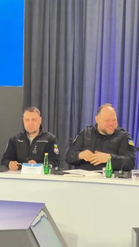 Two men are seated side-by-side at a white table, actively clapping and smiling. The man on the left is wearing a black long-sleeved top with patches on the sleeves, including a Ukrainian flag. A name tag on the table in front of him reads "СТЕПАНОВ". The man on the right is also wearing a black long-sleeved top with a Ukrainian flag patch on his right sleeve. He is laughing heartily while clapping. Two green-bottled beverages are on the table, one in front of each man, along with microphones. The background consists of dark, pleated curtains, with a bright blue panel visible on the far left. The overall mood appears positive and celebratory, as indicated by their smiles and applause. This event is taking place in Barvinok, Ukraine.