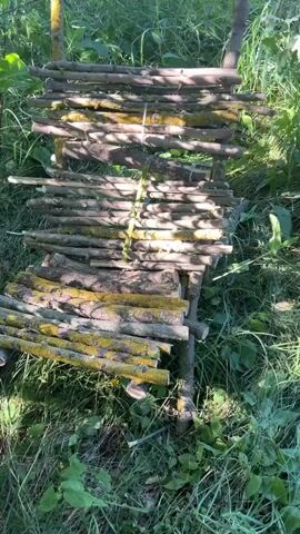 A close-up, low-angle shot captures a makeshift wooden bench constructed from branches and logs, situated outdoors amidst dense green foliage. The bench is built with horizontally placed branches forming the seat and backrest, tied together with thin, light-colored string. Some of the logs and branches are covered in patches of yellow-green lichen. The surrounding environment is overgrown with grass and various green plants, suggesting a natural, uncultivated setting, possibly a forest or garden. The lighting indicates it is daytime, with sunlight casting shadows on the wooden structure. The camera slowly pans across the bench, showcasing its rustic construction and the surrounding vegetation. There are no people visible in the video.