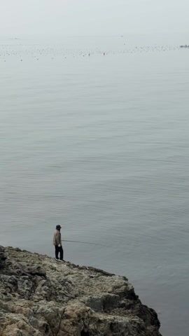 A person stands on a rocky edge overlooking a calm, gray body of water under an overcast sky. The person is wearing a dark hat, a light jacket, dark pants, and white shoes. They hold a fishing rod, suggesting they are fishing. In the distance, there's a faint line of objects, possibly buoys or boats, adding to the muted, somewhat lonely atmosphere of the scene.