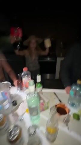 A group of young adults are gathered around a table outdoors at night. A woman in the center, wearing a patterned top and a straw hat, is holding a large bottle of Coca-Cola in one hand and a tan cup in the other, as if making a toast or pouring a drink. A man to her right, wearing a dark shirt, looks off to the side. Another person, mostly out of frame to the left, is holding a red object. The table is cluttered with bottles and cups. A grill is visible in the background. The scene appears to be a casual gathering with drinks.