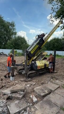 Two men are working at a construction site. One man, wearing an orange shirt and blue shorts, is standing next to a metal post, holding a hammer. The other man, wearing a green hat and plaid shorts, is operating a large yellow drilling machine. The machine has a long, angled boom with a drilling attachment at the top. The site is cleared of debris, with broken concrete pieces scattered around. The background shows trees and a fence under a blue sky with some clouds.