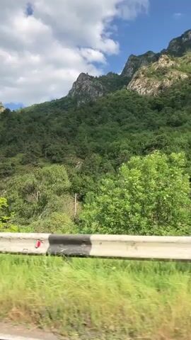 The video shows a view from a moving vehicle, looking out at a lush green mountain landscape on a cloudy day. The foreground is dominated by a white and black striped guardrail. Beyond the guardrail, there are trees and grassy areas. In the background, a large, tree-covered mountain range rises against a blue sky with white clouds. The camera perspective is low, suggesting it is looking out from a car window. There are no people visible in the video. The overall impression is of a scenic drive through a natural environment.