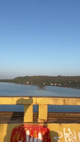 The video shows a view from a bridge over a wide body of water with a green, hilly landmass in the distance. The sky is clear and blue. The bridge's yellow concrete railing has graffiti on it. The road on the bridge is dark asphalt. There are some lamp posts along the side of the road and a few vehicles driving in the distance. The water is relatively calm. The camera pans slightly to the right, revealing more of the bridge and the surrounding landscape. The video seems to be taken from a moving vehicle on the bridge.
