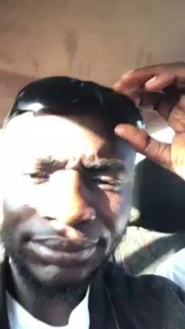 A close-up shot inside of a vehicle shows a Black man wearing a black jacket. He is adjusting a pair of sunglasses that are on his head. He has a short beard and his skin is sweaty. He looks into the camera and mouths words to someone out of frame. A person wearing a white shirt is partially visible in the passenger seat. The interior of the vehicle appears to be tan.