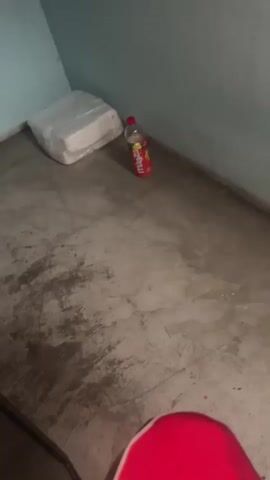 The video shows a corner of a room with a concrete floor and pale blue walls. In the corner, a large white object, possibly a mattress or a bale of something wrapped in plastic, sits on the floor. Next to it, a red plastic bottle with a yellow label is standing upright. The bottle appears to contain a liquid. The camera is positioned low, looking towards the floor, and a portion of a bright pink object, likely clothing, is visible in the bottom foreground. The lighting is dim.