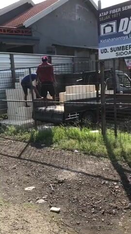 Two men are seen unloading white rectangular objects from the back of a dark-colored truck. One man, wearing a blue shirt and dark pants, is bent over, appearing to lift an object. The other man, in a red shirt and dark shorts, is standing and looking towards the truck. The truck is parked on a dirt surface next to a building with a red roof. There is a metal fence in the foreground and a sign on the building. The environment appears to be outdoors with some vegetation.