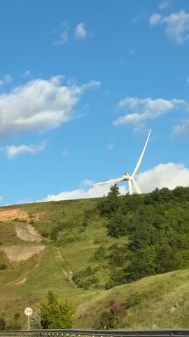 A wind turbine with white blades stands on a grassy hill under a blue sky with white clouds. Below the hill, a road leads to a tunnel. Several vehicles, including vans and cars, are on the road, some approaching the tunnel entrance. A speed limit sign showing "70" is visible on the side of the road. The landscape is characterized by rolling hills covered in green grass and sparse trees.