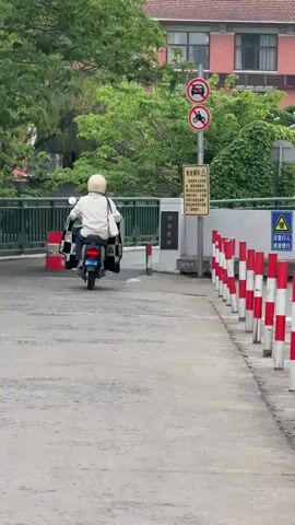 A man wearing a white jacket and a white helmet is riding a black electric scooter away from the camera. He passes a red and white striped barrel, and then drives onto a bridge. A second man riding an electric scooter follows close behind, but in the opposite direction. He is wearing a green shirt, blue helmet, and white shoes. The bridge is gray and has a green metal railing. The area is surrounded by green trees and a few signs.
