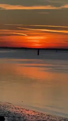 The video shows a tranquil scene of a sunset over the water. The sky is painted with vibrant hues of orange and red, streaked with lines of white clouds. The sun is setting on the horizon, casting a warm, reflective glow across the water's surface. Several dark wooden posts are sticking up in the water in the distance. The foreground features a beach with dark sand. The overall atmosphere is serene and beautiful.