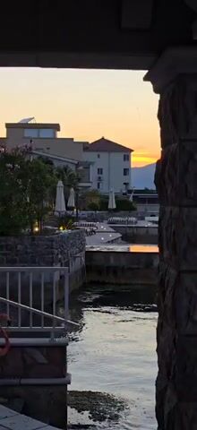 The video pans over a stone-and-concrete patio on the water at dusk. A stream of water pours from a pipe into the water, which reflects the orange sunset. Several empty lounge chairs are arranged on stone tiers leading up the patio. White patio umbrellas are closed and standing nearby. Tiny lights set into the stones provide illumination as well. Buildings can be seen behind the patio.