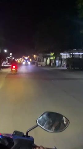 The video, filmed at night from a motorcycle, shows a street scene with a bus approaching. The bus has large, cartoonish eyes painted on its front, resembling the character Lightning McQueen from the movie Cars. The street is dimly lit by streetlights and the headlights of vehicles. A red taillight of another motorcycle is visible in the distance. Trees and buildings line the sides of the street. The camera's perspective is from the rider's point of view, with a motorcycle mirror in the foreground. The bus passes by, its headlights illuminating the road. The video then continues to show the street as the motorcycle moves forward.