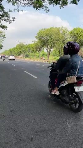 A person is riding a motorcycle on a paved road. The person is wearing a helmet. The motorcycle has a passenger behind them, also wearing a helmet. The passenger is carrying a purse on their shoulder. The road is lined with trees and there are other vehicles in the background. The sky is cloudy.