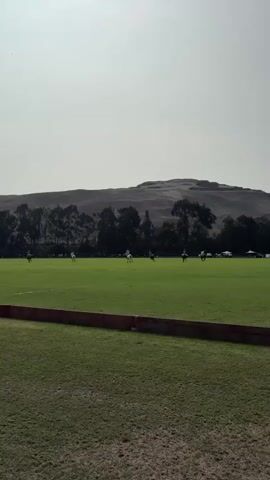 The video shows a polo match from a low angle, with a fence in the foreground. The field is green and expansive, with a line of trees in the background. Beyond the trees is a large, barren hill under a hazy sky. Several polo players on horseback are visible on the field, engaged in the game. Their movements are captured as they ride and swing mallets. The atmosphere appears to be outdoors on a bright day.
