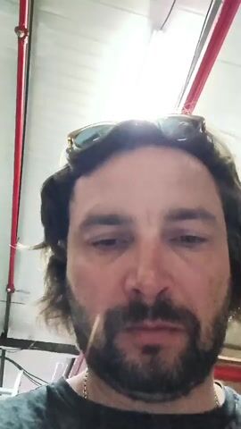 The video shows a close-up, first-person perspective of a man's face. He has brown hair, a beard, and sunglasses perched on his head. He is looking directly at the camera with wide, surprised eyes. He appears to be in an indoor environment with exposed pipes and lighting visible in the background. The man's mouth is slightly open. There is a toothpick or similar small object protruding from his mouth. The lighting in the scene is somewhat harsh, casting shadows on his face.