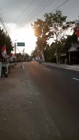 A low-angle, handheld shot of a road in what appears to be Southeast Asia at dusk. The sun is setting, casting a warm glow over the scene. The road is paved and has a white dividing line. On the left side of the road, there are some small market stalls, one of which has a sign that reads "Teh Solo." Indonesian flags are displayed along the roadside. Several vehicles, including a white van, are on the road. A small blue vehicle, possibly a motorcycle or scooter, is also visible. Trees and power lines line the road. The sky is partly cloudy.
