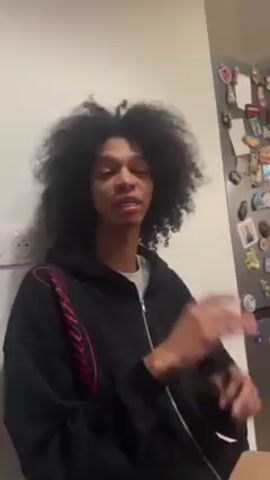 The video shows a person with dark skin and long curly hair. The person is wearing a black hoodie with pink writing on it. They are standing in front of a white wall with an electrical outlet and a refrigerator with papers attached. The person is talking, looking off to the side, and gesturing with their hands. They say "Dancing on different kind of music. Classic. Arabic."