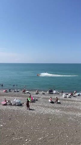 The video shows a sunny day at a pebble beach. The sea is a vibrant turquoise, with gentle waves. Several people are visible on the beach, relaxing on lounge chairs and swimming in the water. In the distance, two jet skis are active on the water. One jet ski is towing a circular inflatable tube with a person riding on it. The second jet ski is being ridden by another person, who is creating a large spray of water as they maneuver. The sky is a clear, bright blue with a few wispy clouds. The overall scene depicts a typical summer day at a beach resort.