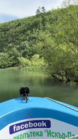 The camera is on a small blue boat on a river, pointing forward. The river is green and the water is rippling. The river is flanked by hills covered in green trees and other foliage. The sky is blue with some white clouds. The boat has a sticker that includes the word "facebook".