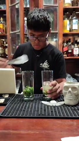 A young man with dark hair and glasses is making a drink at a bar. He is wearing a black t-shirt. He uses tongs to add ice to a glass that already contains mint and a clear liquid. Another glass beside it contains only mint. In front of him is a black rubber mat on a wooden bar. A skull-shaped cup and a spoon are to the right. Shelves stocked with bottles are visible behind him.