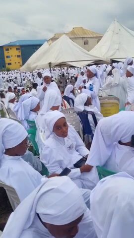 The video shows a large outdoor gathering of people dressed in white robes and head coverings, possibly attending a religious event. The crowd is seated in white plastic chairs on a grassy field, with tents set up in the background. A few buildings are also visible in the distance. The people appear to be of African descent. Some individuals are interacting with each other, while others are looking towards the front of the gathering. At least one person is raising their arms in what could be a gesture of worship or celebration.
