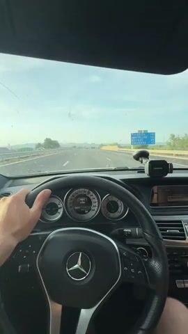 A first-person perspective video shows a person driving a Mercedes-Benz car on a highway. The steering wheel and dashboard are visible, with the driver's hand on the wheel. The car appears to be moving at a moderate speed. The road ahead is clear, with other vehicles in the distance. The sky is bright and cloudy. The video appears to be taken during the daytime.