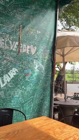 The video shows the outdoor seating area of a restaurant or bar. The area is partially covered, with a chalkboard-like wall covered in signatures and messages. There are several tables and chairs, some under umbrellas, made from rustic materials like wood and recycled cable spools. In the background, there are picnic tables with attached swings, set on a gravel surface. Beyond the seating area, there's a glimpse of a rural landscape with green fields. A stone building is also visible, adding to the rustic charm. The sound of music can be heard in the background.