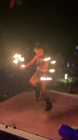 A young woman performs a fire dance on a stage at night. She is wearing a black leather-style bra, belt, shorts, fishnet stockings, and boots. She spins and twirls a pair of implements that each contain multiple flames. The stage is painted with concentric circles. There are people sitting at tables and chairs in the background, and small glowing lights on the tables. The video is shot at a low angle.