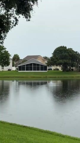 The video shows a lake with ripples on the surface. There is a screened-in building on the opposite shore, behind which are houses and trees. The sky is overcast, and it appears to be raining. The foreground shows a grassy embankment.