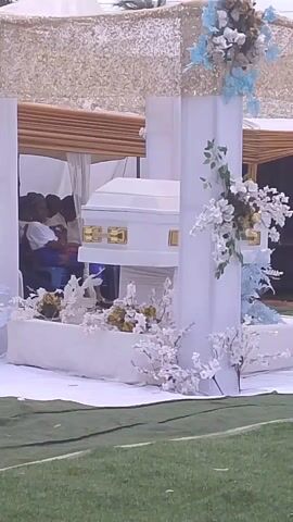 The video shows a white coffin with gold handles under a canopy decorated with white and blue flowers. The coffin is placed on a raised platform covered in white cloth, surrounded by additional floral arrangements. People are seated in the background under the canopy. The scene is set outdoors on a green grassy area.