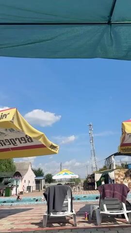The video opens with a view of a clear blue sky and some white clouds. In the foreground, a yellow umbrella is partially visible on the left side, and two white lounge chairs with towels are in the center. The pool area is in the background, with several people swimming and enjoying themselves. A cell tower stands tall in the distance, and a few buildings and trees are scattered around the perimeter of the pool. The overall scene depicts a relaxed and sunny day at an outdoor pool.