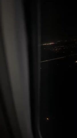 The video shows a night view from an airplane window. City lights are visible in the distance, appearing as scattered points and lines of light against the dark background. Some light trails are also visible, potentially from ground traffic. The interior of the airplane is dimly visible, with the window frame partially obscuring the view.