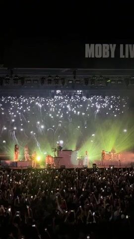 A wide shot captures a large outdoor concert audience with their cell phones held up, illuminating the space. On stage, a band performs under bright lights, with a large screen behind them displaying a black and white image of a woman singing. The text "MOBY LIVE" is visible in large white letters across the top of the screen.
